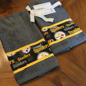 May include: Two gray kitchen towels with a yellow and black Steelers logo and the word "Steelers" repeated on the border.