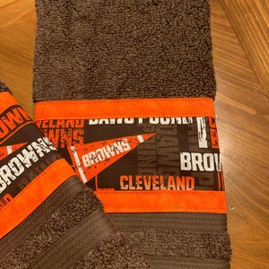 May include: Two brown towels with orange trim. The towels have a Cleveland Browns theme with the words "Cleveland Browns" printed on them.