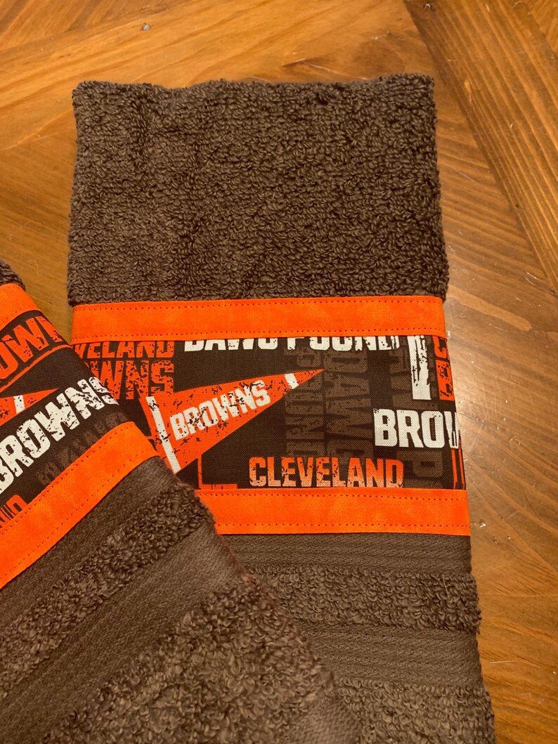 May include: Two brown towels with orange trim. The towels have a Cleveland Browns theme with the words "Cleveland Browns" printed on them.