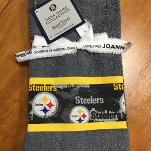 May include: Gray hand towel with a yellow border and a black and white pattern of the Pittsburgh Steelers logo. The towel is labeled "Sara Home Collection" and "Designed in Hudson, Ohio Exclusively for Joann".
