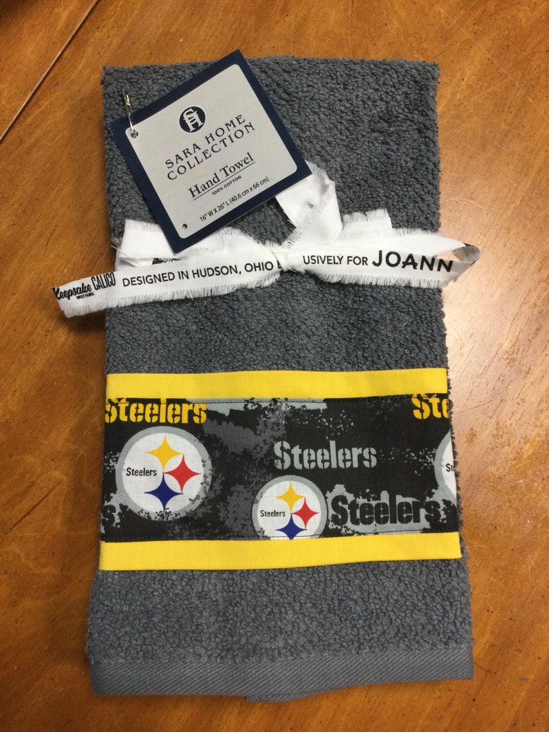 May include: Gray hand towel with a yellow border and a black and white pattern of the Pittsburgh Steelers logo. The towel is labeled "Sara Home Collection" and "Designed in Hudson, Ohio Exclusively for Joann".