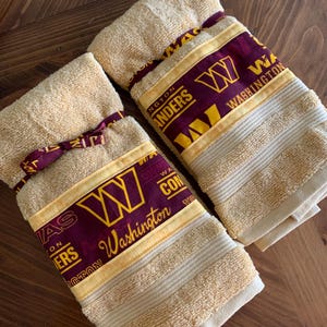 May include: Two tan hand towels with a burgundy and gold Washington Huskies design. The towels have a yellow ribbon tied around them.