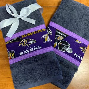 May include: Two dark gray hand towels with a purple and gold Baltimore Ravens themed border. The towels are tied together with a white ribbon.