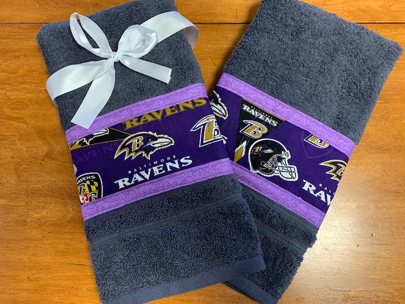 May include: Two dark gray hand towels with a purple and gold Baltimore Ravens themed border. The towels are tied together with a white ribbon.