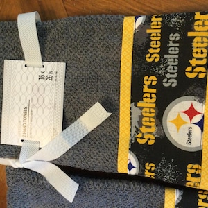 May include: Two gray hand towels with a black and yellow Steelers logo and the words "Steelers" repeated on the fabric. The towels are 16 x 26 inches.