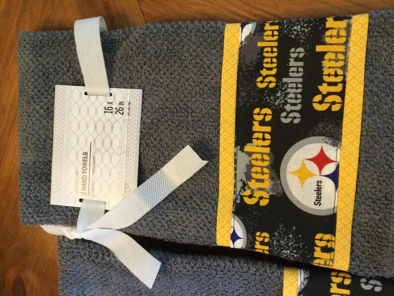 May include: Two gray hand towels with a black and yellow Steelers logo and the words "Steelers" repeated on the fabric. The towels are 16 x 26 inches.