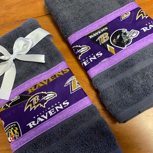 May include: Two gray hand towels with purple and gold trim featuring the Baltimore Ravens logo and team name. The towels are tied with a white ribbon.