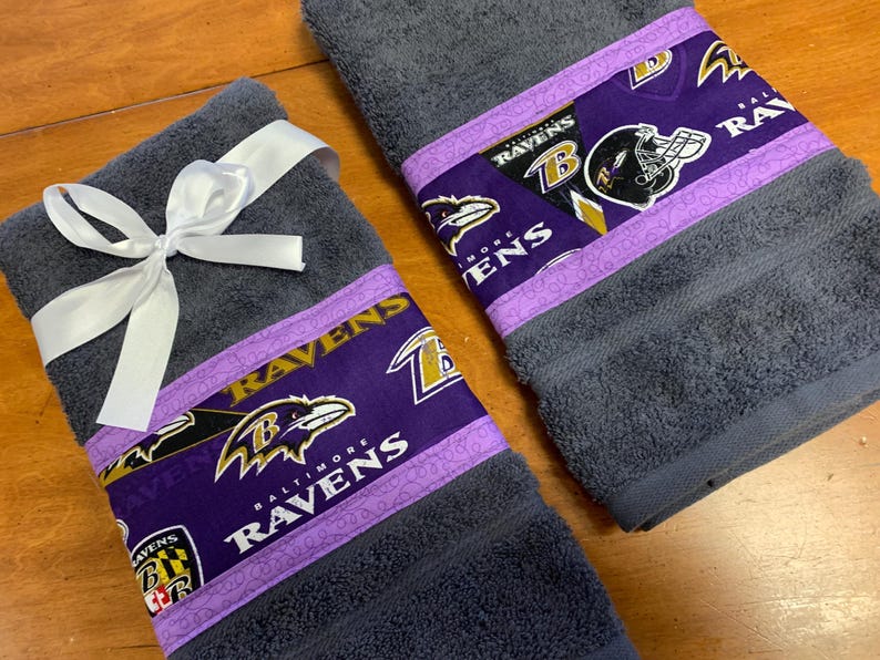 May include: Two gray hand towels with purple and gold trim featuring the Baltimore Ravens logo and team name. The towels are tied with a white ribbon.