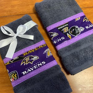 May include: Two gray hand towels with purple and gold trim featuring the Baltimore Ravens logo and the words "Baltimore Ravens". One towel is tied with a white ribbon.