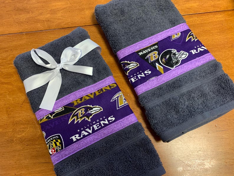 May include: Two gray hand towels with purple and gold trim featuring the Baltimore Ravens logo and the words "Baltimore Ravens". One towel is tied with a white ribbon.