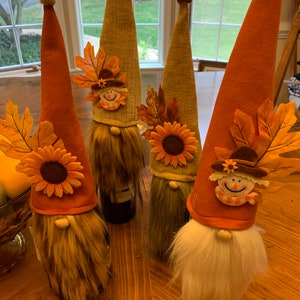 May include: Four fall-themed gnome bottle stoppers with orange, brown, and beige hats. Each gnome has a white beard and a sunflower with brown leaves on their hat. The gnomes are sitting on a wooden table.
