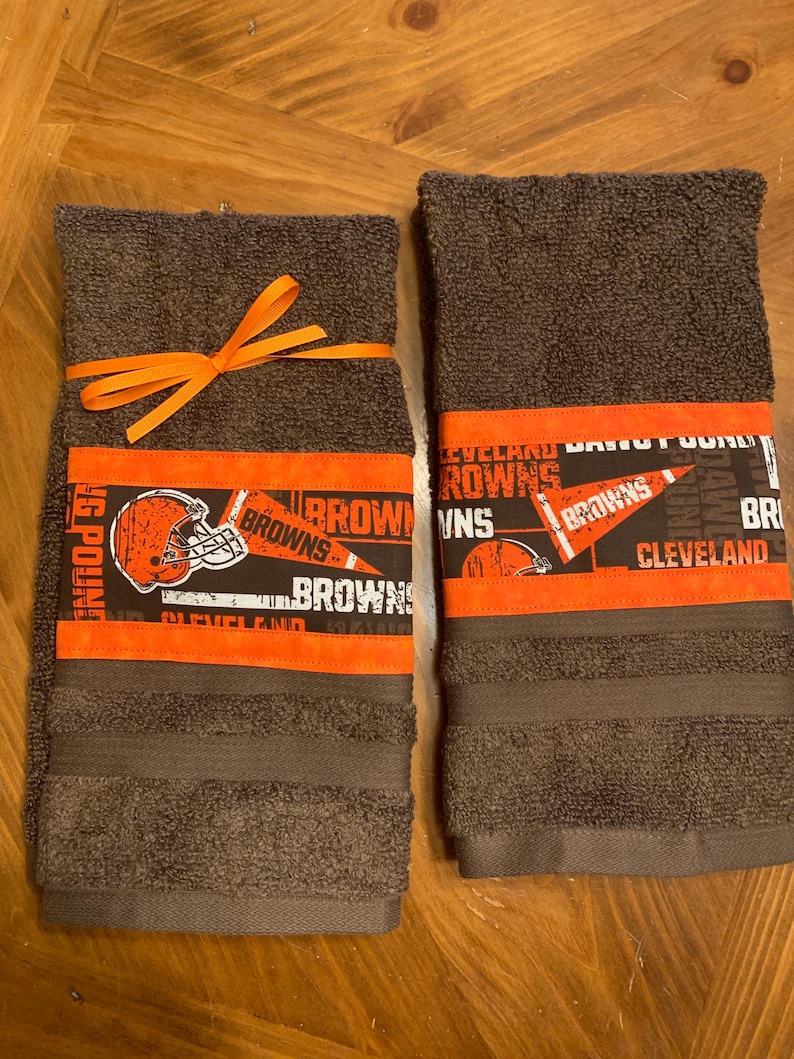May include: Two brown hand towels with orange and black Cleveland Browns themed fabric trim. The towels are tied together with an orange ribbon.