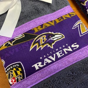 May include: Purple and black towel with a Baltimore Ravens logo and the words "Baltimore Ravens" on it. The towel has a purple and white trim with a pattern of swirls.