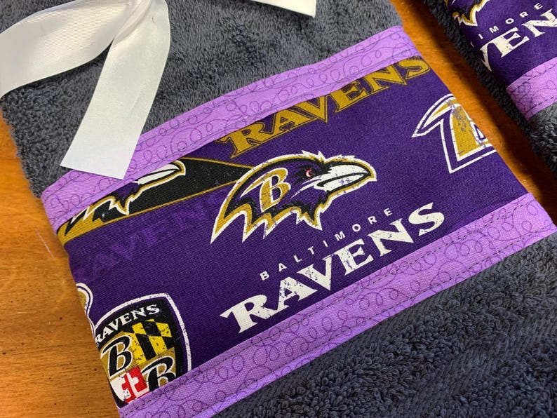 May include: Purple and black towel with a Baltimore Ravens logo and the words "Baltimore Ravens" on it. The towel has a purple and white trim with a pattern of swirls.