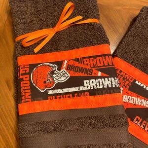 May include: Two brown kitchen towels with orange trim and a Cleveland Browns football team design. The towels are tied together with an orange ribbon.