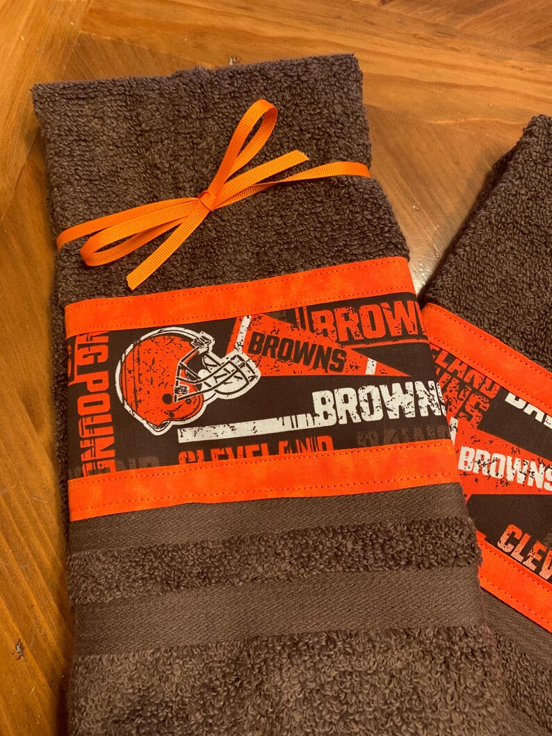 May include: Two brown kitchen towels with orange trim and a Cleveland Browns football team design. The towels are tied together with an orange ribbon.