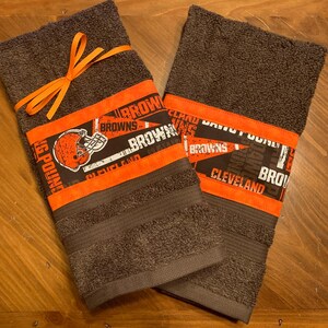 May include: Two brown towels with orange and white trim featuring the Cleveland Browns logo and text. The towels are tied together with an orange ribbon.