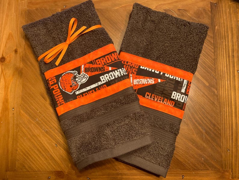 May include: Two brown towels with orange and white trim featuring the Cleveland Browns logo and text. The towels are tied together with an orange ribbon.