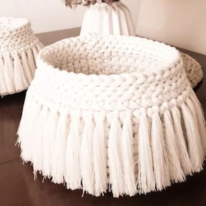 Peut inclure: Panier en macramé blanc avec un motif tissé et une bordure à franges. Le panier est rond et possède une large ouverture. Le panier est fait d'un matériau épais et doux, probablement pour le rangement ou la décoration.