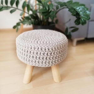 Könnte beinhalten: Ein runder Hocker aus hellgrauem Häkelgarn mit drei Holzbeinen. Der Hocker ist rund und hat eine strukturierte Oberfläche.