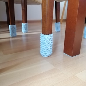 Op de afbeelding: Close-up van vier lichtblauwe gebreide stoelpootbeschermers die hardhouten vloeren beschermen. De beschermers zijn gemaakt van een zacht, getextureerd garen en zijn ontworpen om krassen en geluid te voorkomen.