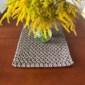 Könnte beinhalten: Ein quadratischer, taupefarbener Makramee-Tischläufer mit strukturiertem Muster. Eine klare Glasvase enthält einen Strauß leuchtend gelber Blumen, wodurch ein Kontrast von Farben und Texturen entsteht. Der Tischläufer liegt auf einer braunen Holzoberfläche.