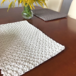 Puede incluir: Dos manteles individuales de crochet, uno blanco y otro gris, sobre una mesa de madera. El mantel individual blanco está en foco y tiene un diseño texturizado y tejido.