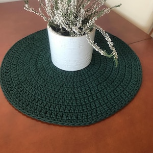 Op de afbeelding: Een ronde, donkergroene gehaakte tafelmat met een witte keramische pot en witte bloemen erop.