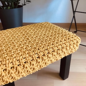Op de afbeelding: Een gele gehaakte kruk met zwarte houten poten.