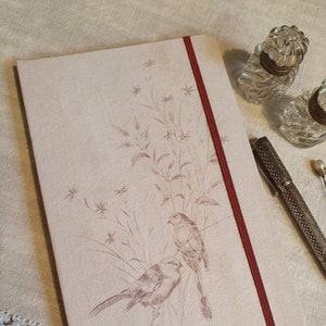 Może przedstawiać: Beżowy notes z lnu z czerwonym zapięciem wstążką. Okładka przedstawia delikatną ilustrację dwóch ptaków siedzących na gałęzi z liśćmi i kwiatami.