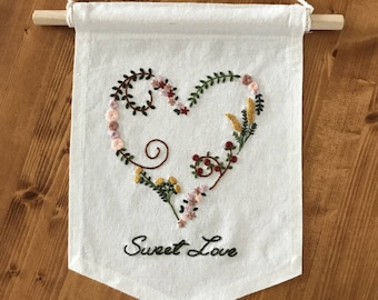 Könnte beinhalten: Weißes Banner mit einer Holzstange und einem Seilaufhänger. Das Banner zeigt ein gesticktes Herz, das mit bunten Blumen und Grün gefüllt ist. Die Worte "Sweet Love" sind darunter gestickt.