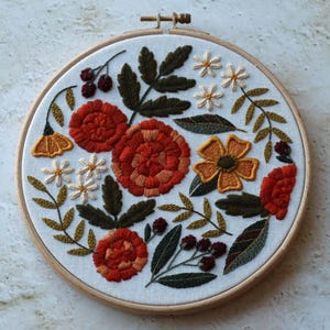 Peut inclure: Broderie florale faite à la main dans un cercle de bois. Le motif présente des fleurs orange, jaunes et blanches vibrantes, avec des feuilles et des baies vertes. La broderie est réalisée sur un tissu blanc, créant une œuvre d'art colorée et texturée.