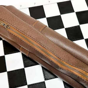 May include: A brown leather chess set roll with a zipper closure. The roll is lying on a white and black checkered chessboard.
