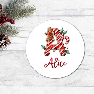 Könnte beinhalten: Ein weißer runder Anhänger mit einem roten und weißen Zuckerstangenbuchstaben A, einem Lebkuchenmann und Stechpalmenblättern. Der Name "Alice" ist in roter Kursivschrift unter dem Buchstaben geschrieben.