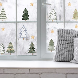 May include: A window with a white frame decorated with green, white, and blue watercolour Christmas trees and gold stars. The window is decorated with a grey knitted cushion and a white cushion with the text "Let it Snow".