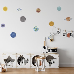 May include: A white wall with a variety of colorful planets and stars, including a sun, a moon, and a ringed planet. There is a white wooden shelf with a star, a whale, and a panda cutout. The shelf is decorated with a toy guitar, a drum, and a small stool. The wall also has a string of wooden clothespins with letters that spell out "HURS".