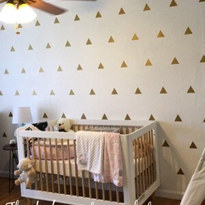 May include: A white cot with a wooden frame and a white and pink blanket inside. The wall behind the cot is decorated with gold triangle stickers. A white teepee tent is in the corner of the room.