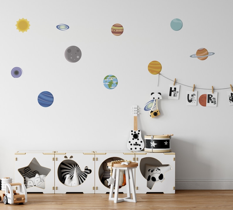 May include: A white wall with a variety of colorful planets and stars, including a sun, a moon, and a ringed planet. There is a white wooden shelf with a star, a whale, and a panda cutout. The shelf is decorated with a toy guitar, a drum, and a small stool. The wall also has a string of wooden clothespins with letters that spell out "HURS".