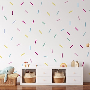 Könnte beinhalten: Eine weiße Wand mit bunten Streusel-Aufklebern, eine weiße Kommode mit zwei Schubladen und Weidenkörben, ein blauer Plüschwal und eine Plüschgiraffe.