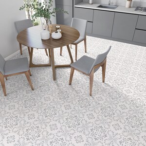 Op de afbeelding: Een houten eettafel met vier grijze stoelen, geplaatst op een witte vloer met een grijs bloemenpatroon. De tafel heeft een houten blad en een grijs onderstel.
