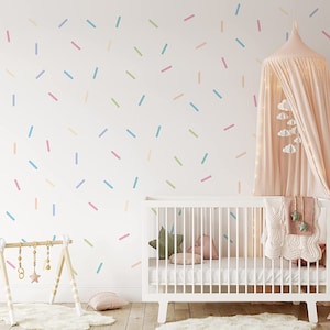 Könnte beinhalten: Ein weißes Babybett mit einer rosa-weißen Decke und einem rosa Baldachin mit weißen Wolken, die daran hängen. Die Wand hinter dem Bett ist mit bunten Streuseln verziert.