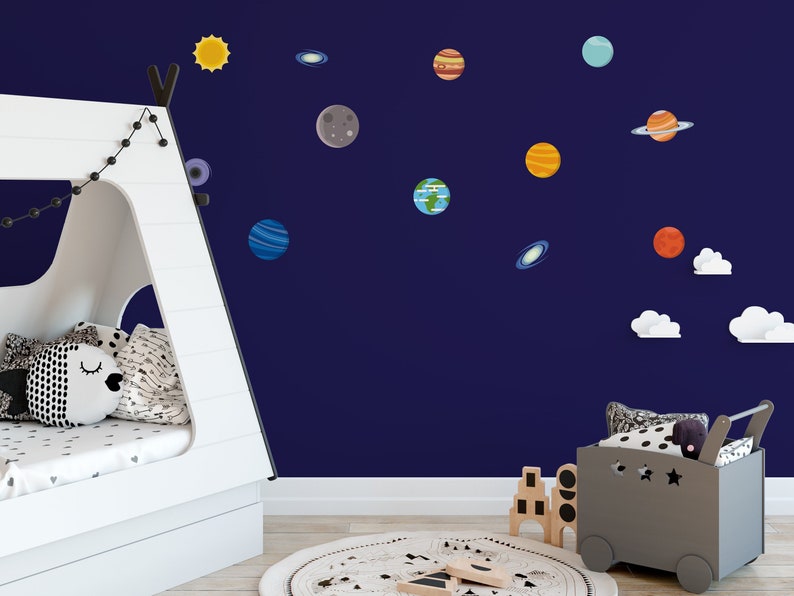 May include: A white teepee tent with a string of white lights hanging from the top. The tent is in a room with a blue wall decorated with planets and stars. There is a gray toy wagon with a black and white polka dot pillow and a black stuffed animal in it.