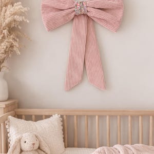 Pode incluir: Uma grande decoração de parede em forma de laço de veludo cotelê rosa empoeirado, com um nó central de padrão floral colorido. O laço tem duas pontas longas e pendentes e uma superfície de tecido canelado, montado sobre um berço de madeira.