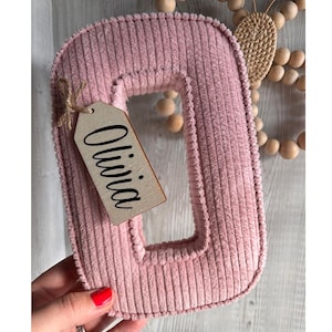 Puede incluir: Una letra "O" de pana rosa, con una etiqueta de madera que dice "Olivia" en escritura negra. La letra está sujeta por una mano con esmalte de uñas rojo. Cuentas de madera decorativas y un elemento tejido en el fondo.