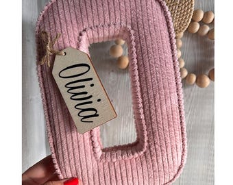 Puede incluir: Una letra "O" de pana rosa, con una etiqueta de madera que dice "Olivia" en escritura negra. La letra está sujeta por una mano con esmalte de uñas rojo. Cuentas de madera decorativas y un elemento tejido en el fondo.