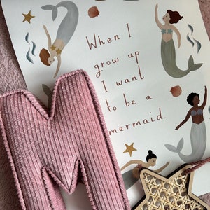 May include: A pink corduroy letter "M" rests against a print featuring mermaids, seashells, and stars. The print reads "When I grow up I want to be a mermaid." A wooden star with a woven center is also visible.
