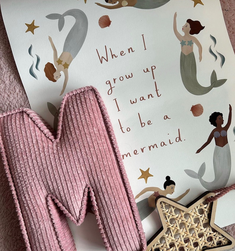 May include: A pink corduroy letter "M" rests against a print featuring mermaids, seashells, and stars. The print reads "When I grow up I want to be a mermaid." A wooden star with a woven center is also visible.