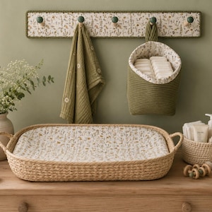 Puede incluir: Un cambiador en cesta trenzada rectangular con forro de estampado floral, un perchero de pared con tela floral, una bolsa de almacenamiento colgante verde oliva, una toalla de muselina verde oliva, una cesta crema con art&iacute;culos de aseo y un peluche.