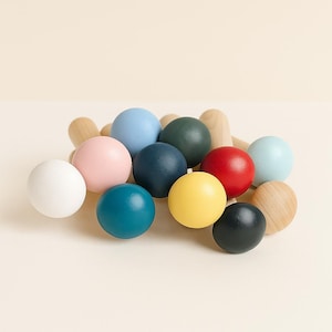 Puede incluir: Una colecci&oacute;n de clavijas de madera con cabezas esf&eacute;ricas pintadas en varios colores, incluyendo blanco, rosa claro, azul claro, azul oscuro, verde oscuro, rojo, amarillo, azul verdoso y negro, junto a varias esferas de madera natural.