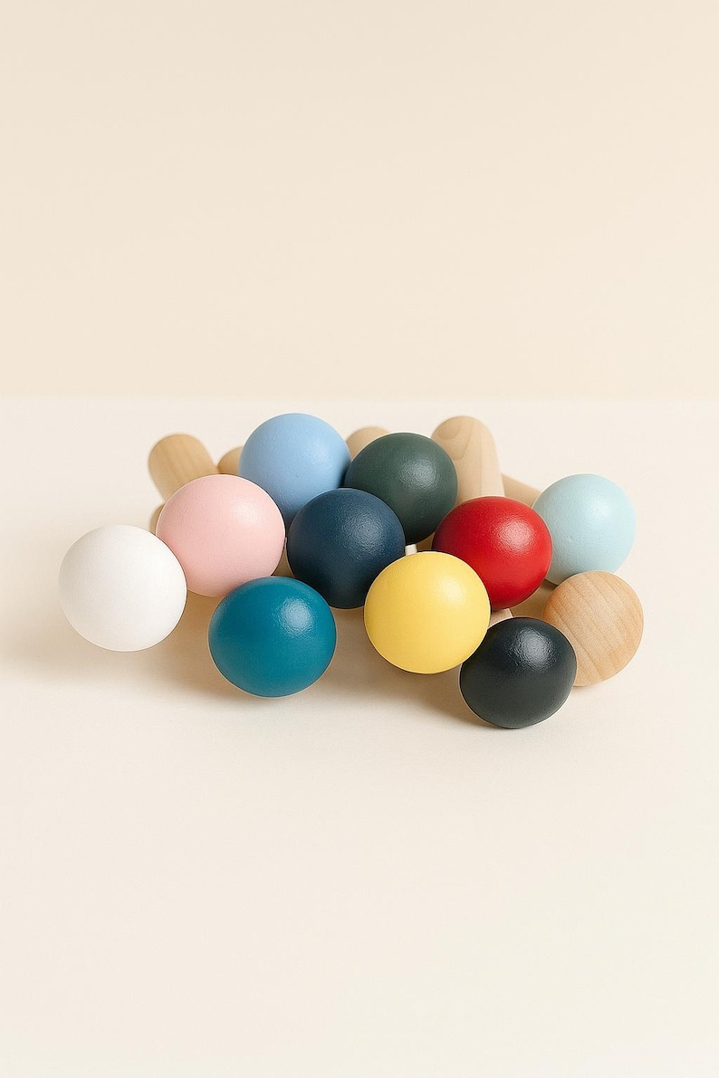 Puede incluir: Una colecci&oacute;n de clavijas de madera con cabezas esf&eacute;ricas pintadas en varios colores, incluyendo blanco, rosa claro, azul claro, azul oscuro, verde oscuro, rojo, amarillo, azul verdoso y negro, junto a varias esferas de madera natural.